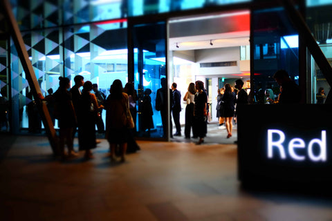 Exterior of Red dot design museum showing the glass facade during night time with groups of people gathering both indoors and outdoors for an event