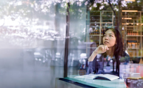 Woman sitting at a table with a book, looking out of a window with a blurred Marina bay background