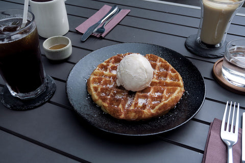 Outdoor seating setting along Marina bay with golden brown waffle with gelato and coconut flakes on a black plate with ice and hot coffee