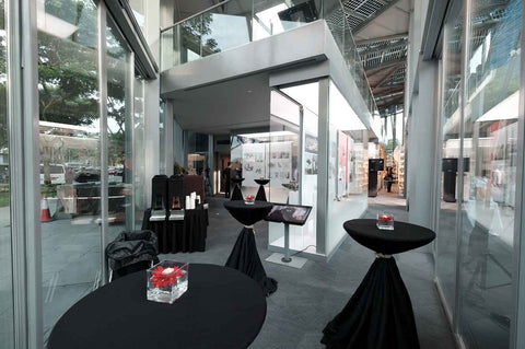 Interior of the gallery exhibit with glass panels, black cocktail tables, and a refreshment counter overlooking Marina Bay