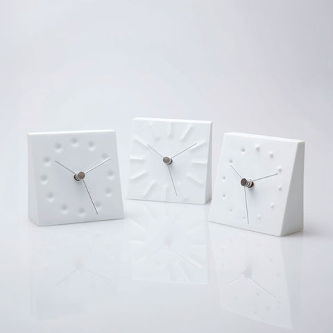 Three minimalist white square wall clocks with textured geometric faces and slim silver hands, arranged on a reflective white surface
