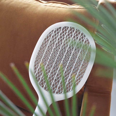 Lumena white insect swatter held in front of a brown couch with green leaves in the foreground