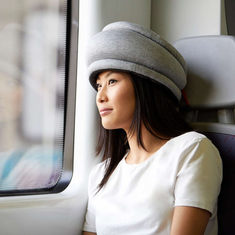 Model relaxes by the window wearing Ostrichpillow Light Versatile Pillow-Midnight Grey, a soft travel companion for comfortable rest on the go