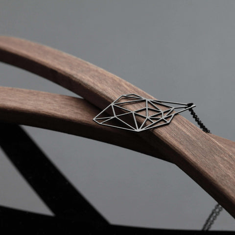 Stainless Steel Pendant - Stone S rests on wood, its sharp geometric lines contrasting warm tones in a clean, minimalist composition