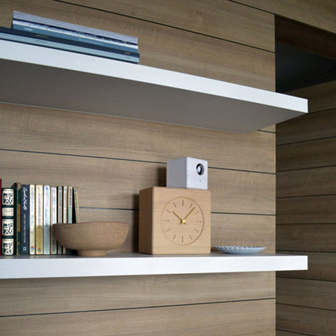 Wooden Cubist Cuckoo Clock with gold hands on a white floating shelf, styled with books, a bowl, and a small white cube.