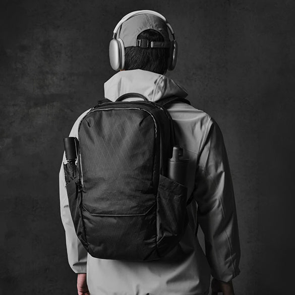 A person viewed from behind wearing a black Alpaka Elements Backpack Pro VX42 over a light gray hooded jacket, with over-ear headphones on, against a dark textured studio background.