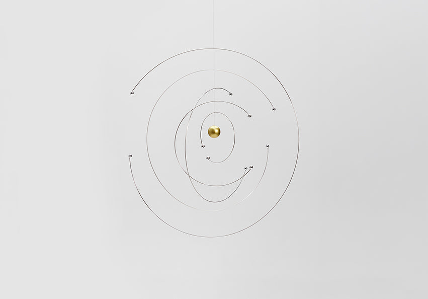 A minimalist hanging mobile with a small gold sphere at the center, suspended against a plain light grey background.
