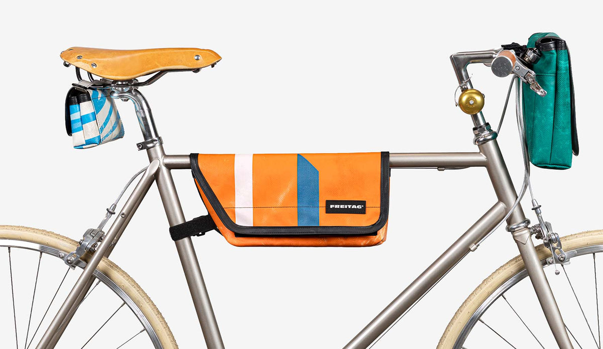 An orange bicycle frame bag with blue and white vertical stripes and a black FREITAG logo, mounted on the downtube of a silver bicycle against a light gray background.