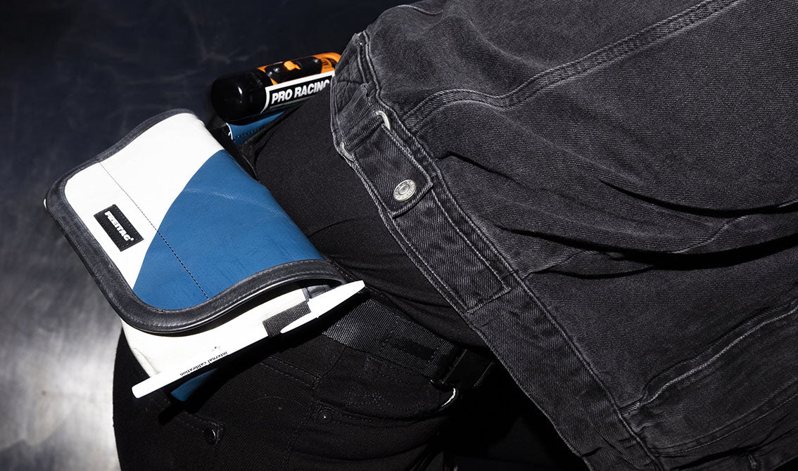 A close-up of a person wearing a black denim jacket and black pants, with a blue and white  Freitag Jamie hipbag attached at the hip.