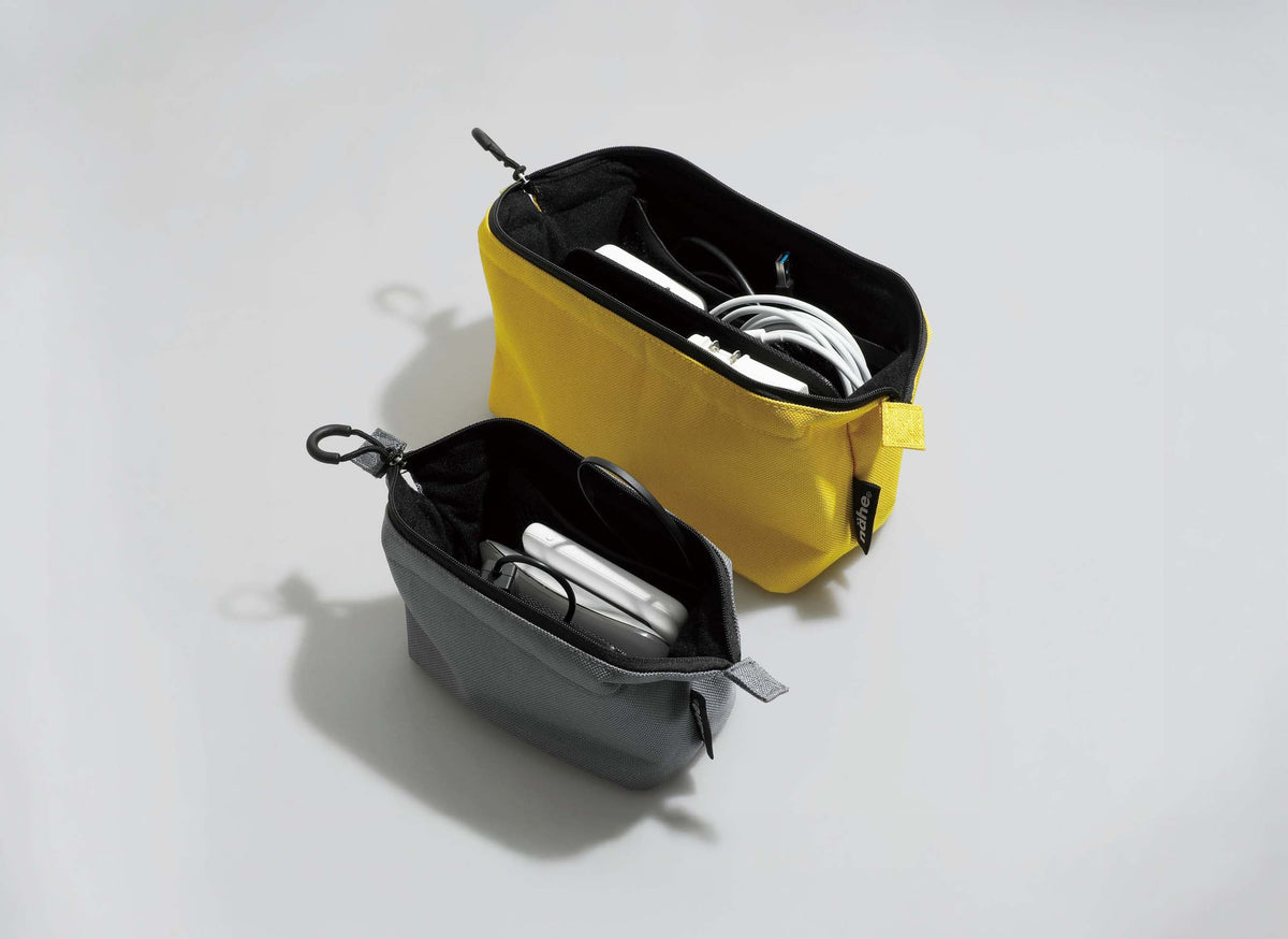 Two HIGHTIDE soft gadget pouches shown open, filled with white cables and silver accessories. The pouch in the back is bright yellow with a black interior, and the pouch in the front is dark gray with a black interior.