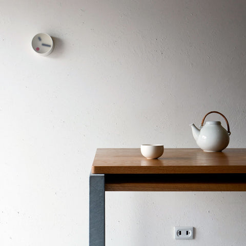 Kehai round wall clock on a textured white wall, with a white ceramic teapot and cup on a wooden table below.