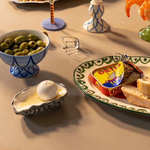 Tablescape with olives in a footed bowl, an oyster-shaped salt shaker, a tin of Ortiz sardines with bread on a patterned plate, and candlesticks.
