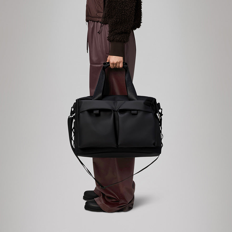 A cropped view of a person wearing burgundy pants holding a Rains Otaru Weekend Bag Small W3 with dual top handles and a detachable shoulder strap