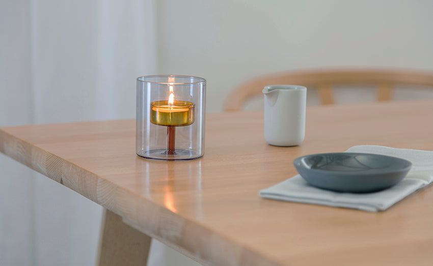 A clear cylindrical, duo-colour glass candle holder containing a lit tealight candle, placed on a light wood table in a softly blurred indoor setting with a small white pitcher and a grey bowl nearby.