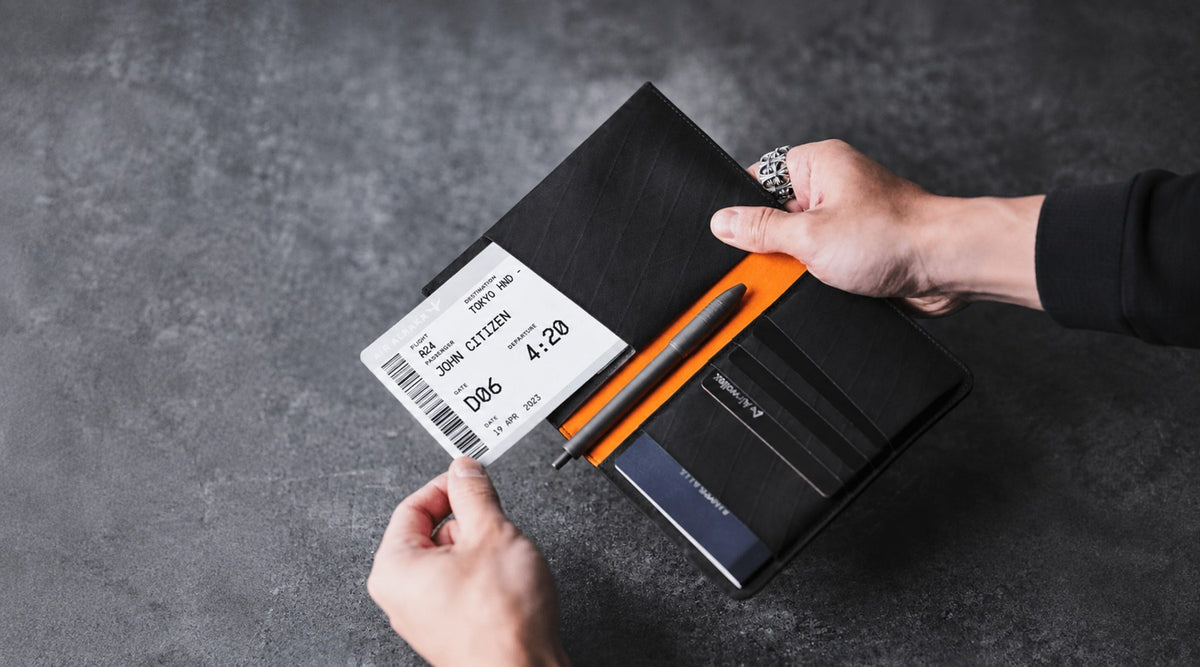 A black Alpaka Ark Bifold Passport Wallet with a boarding pass, pen, card and passport in its sleeve, held by two hands over a dark grey textured surface. 
