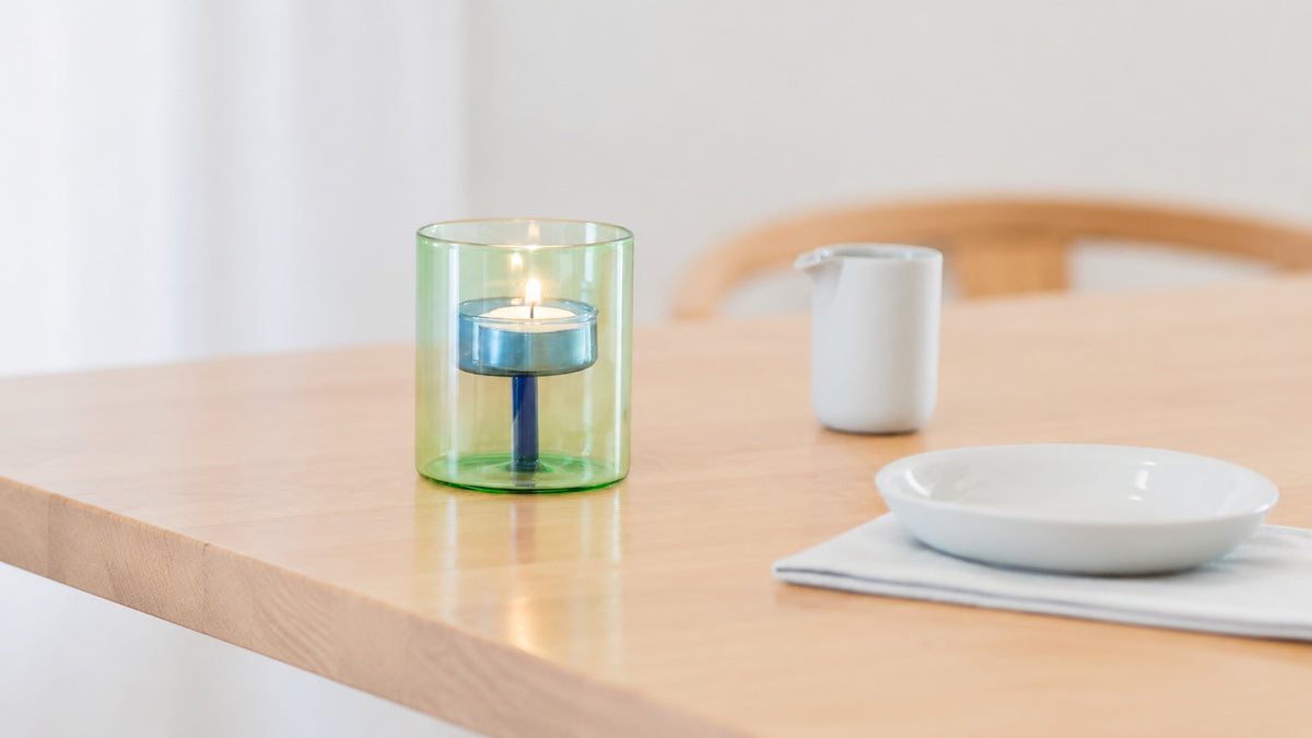 A lit Block Design Duo Tone tealight candle holder in blue and green glass sits on a light wood table, with a white plate and small container blurred in the background.