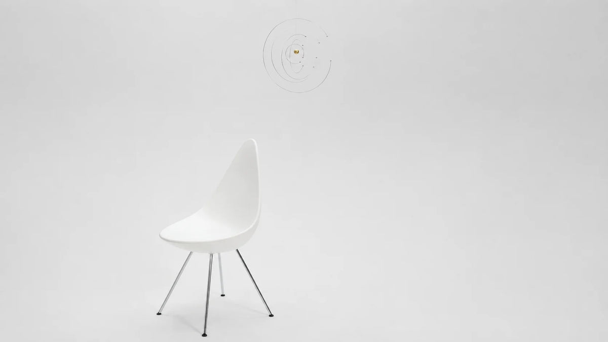 A modern white molded plastic side chair with slim metal legs, set against a seamless light gray studio background. A Flensted Niels Bohrs Atom hanging mobile is hang above and behind the chair.
