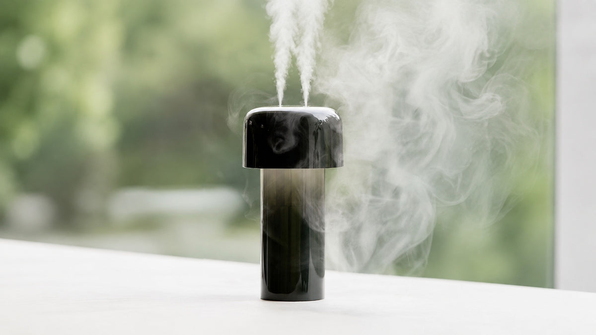 A dark Lumena Mist Stand cylindrical humidifier with two fine mist nozzles spraying vapor, set on a white surface with a blurred green outdoor background.