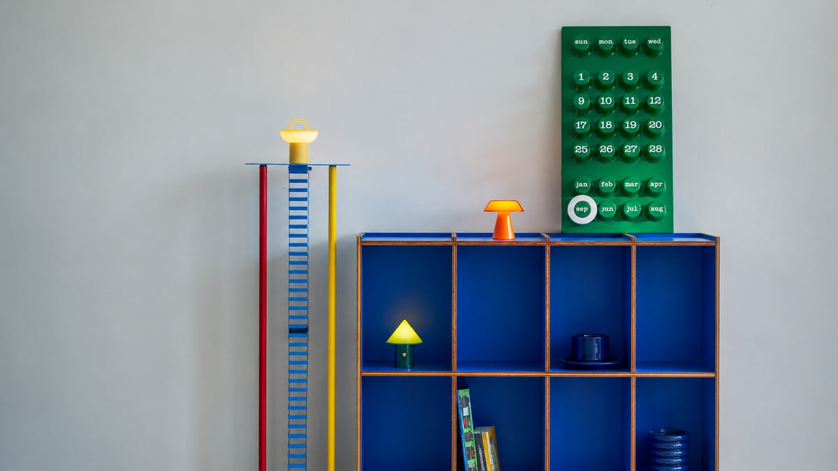 A blue shelving unit with wooden frames, styled with colourful decorative objects including a green calendar, a red, yellow and blue ladder, and small Lumena Space S lamps, set against a plain light gray wall.