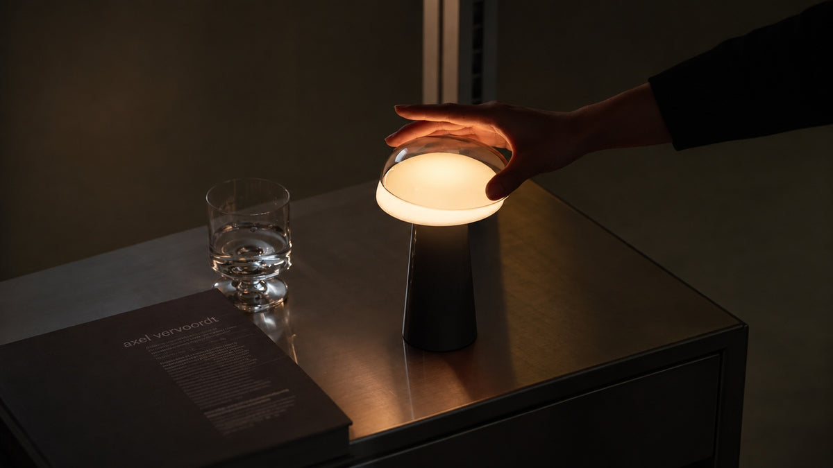 A Lumena Space M1 table lamp with a glowing round shade and dark cylindrical base sits on a dark tabletop, with a hand placing a round light diffuser filter on it. A glass and a dark book are also visible in the dim room setting.