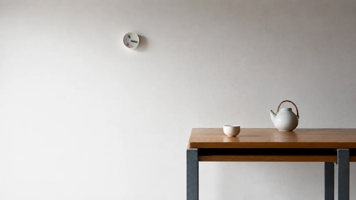 Lemnos kehai round clock hang on a wall behind a brown table that has a teapot and teacup set on it