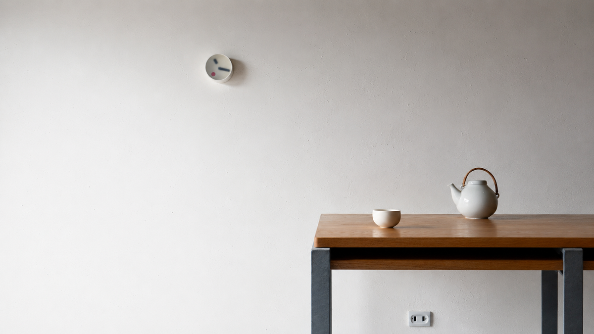 Lemnos kehai round clock hang on a wall behind a brown table that has a teapot and teacup set on it