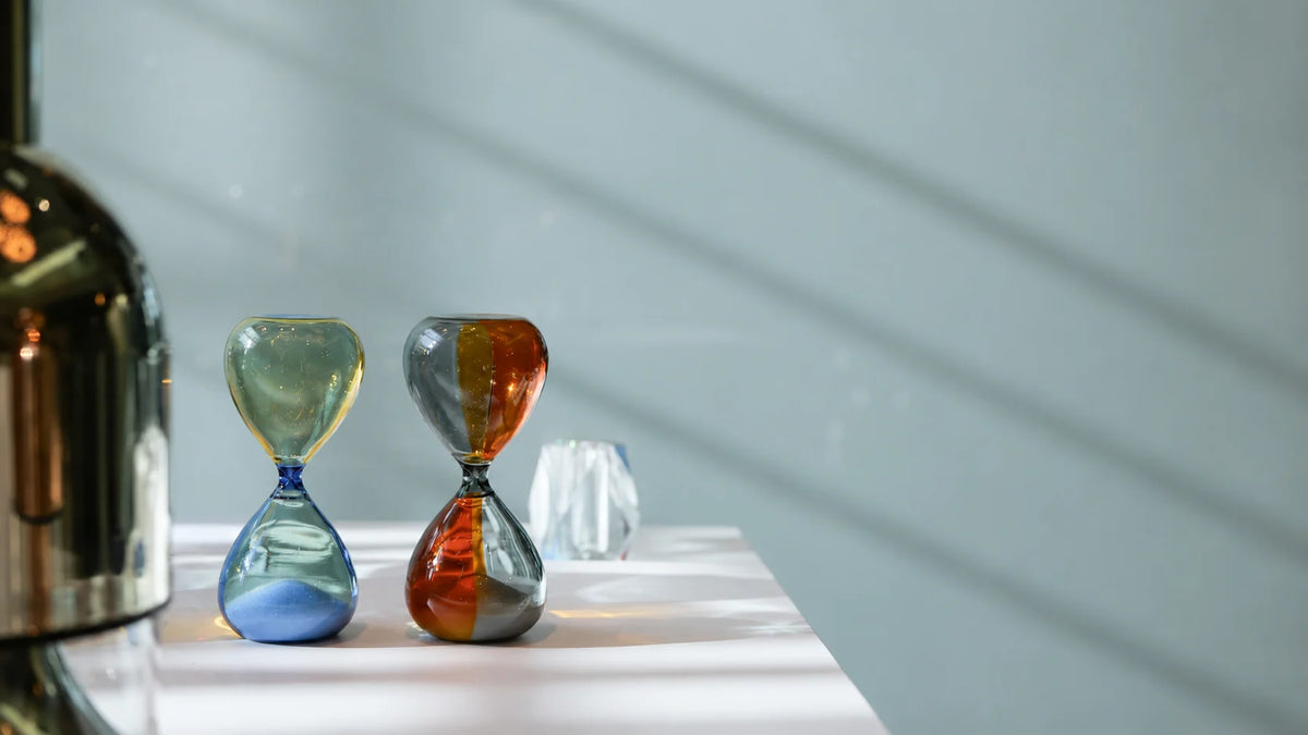Two decorative Hightide Bi-colour hourglasses with blue and yellow, orange and grey patterns sit on a white table next to a large metallic object, with soft diagonal shadows cast across a light blue-gray wall.