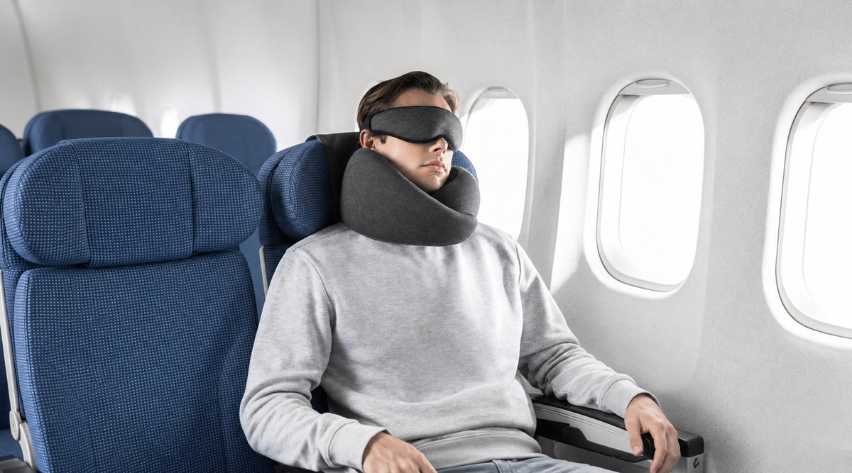 A man wearing a dark Ostrichpillow grey sleep mask and Ostrichpillow Go neck pillow sits in a blue airplane seat by a window.