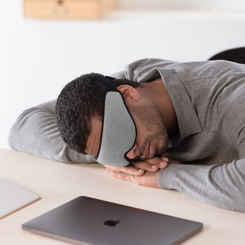 Ostrich Pillow Eye Mask in midnight grey worn by a man napping face-down on a desk beside a laptop.