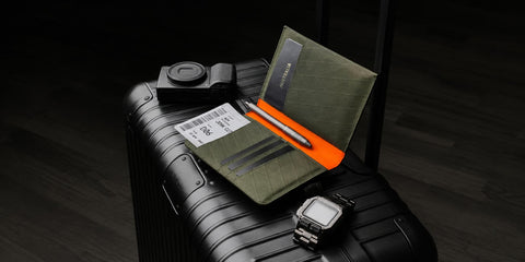 An olive green Alpaka passport holder wallet with an orange internal fabric holding a silver pen, displayed on top of a black hard-shell suitcase alongside a camera, luggage tag, and watch.